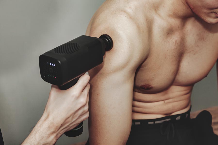 Close up gun percussion massager of shoulder muscle athletic man in medical room at gym. Masseur massage exercises. Percussions therapy for regenerating sport body and rehabilitation injuries