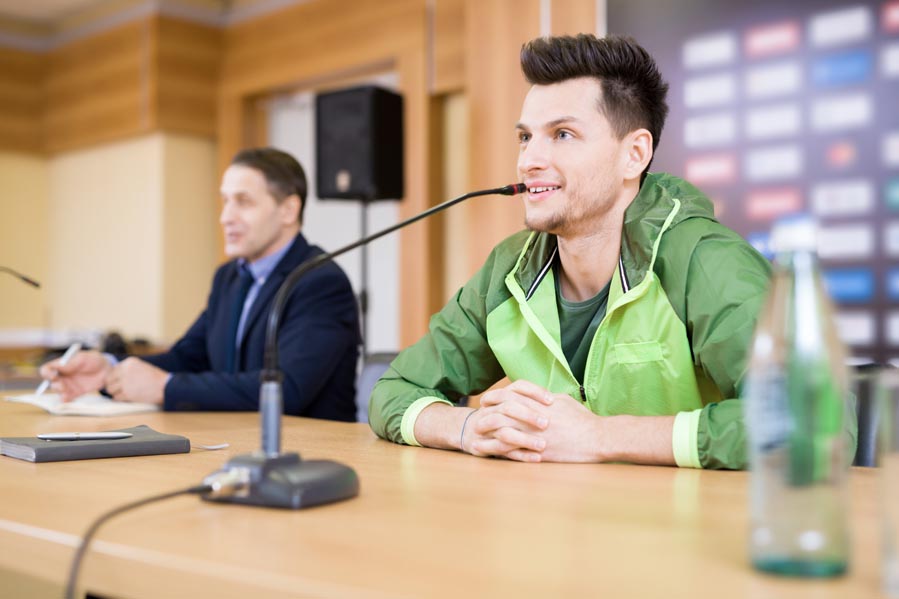 Handsome young football player wearing track jacket expressing his point of view concerning played match while participating in press conference, waist-up portrait